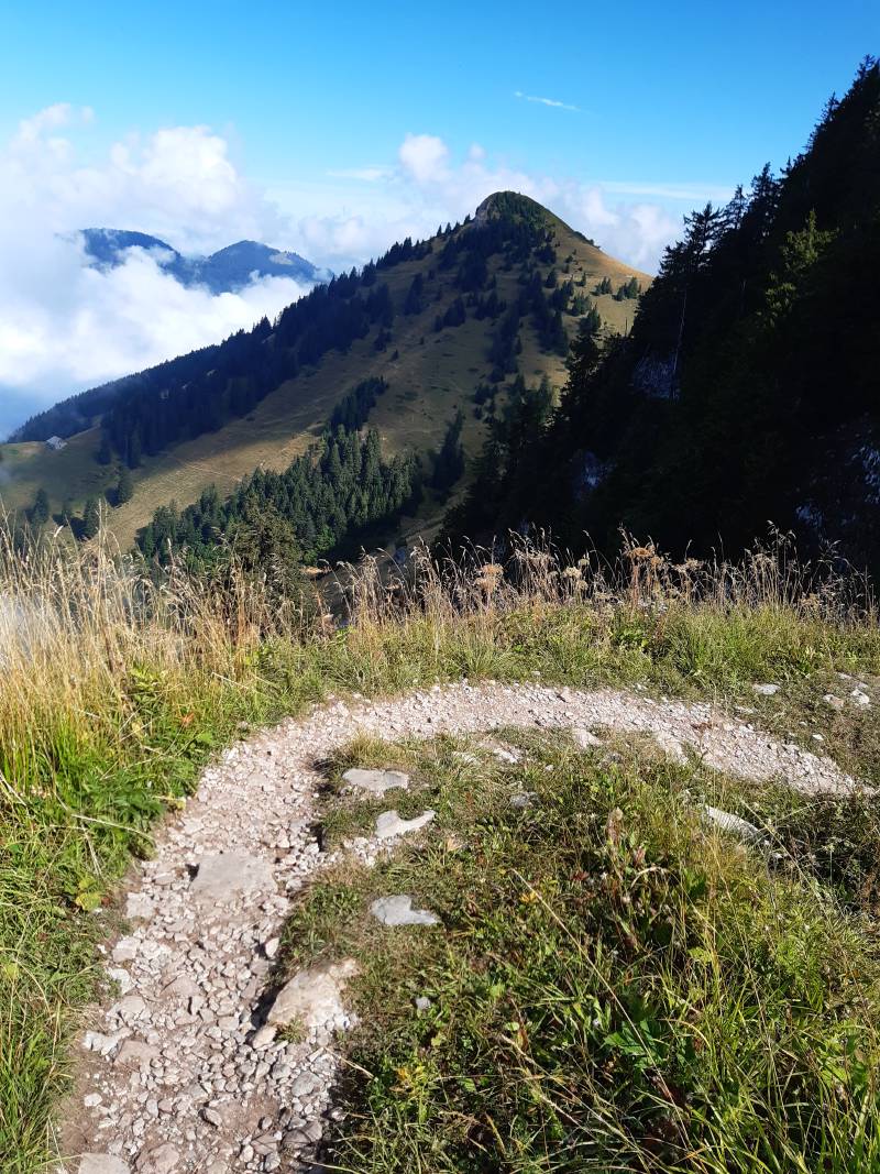 Petit chemin étroit faisant une boucle à 180°; en arrière-plan, un sommet assez proche, et des nuages cachant la vue vers d’autres sommets plus lointains.