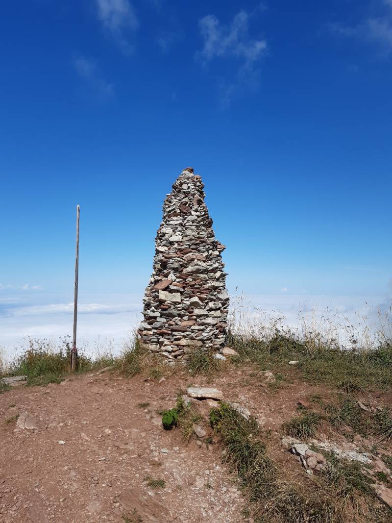 Sur une plateforme de terre avec un peu de verdure s’érige une pyramide faite de pierres de diverses formes et couleurs; en arrière-plan les nuages de basse altitude bouchent complètement la vue.