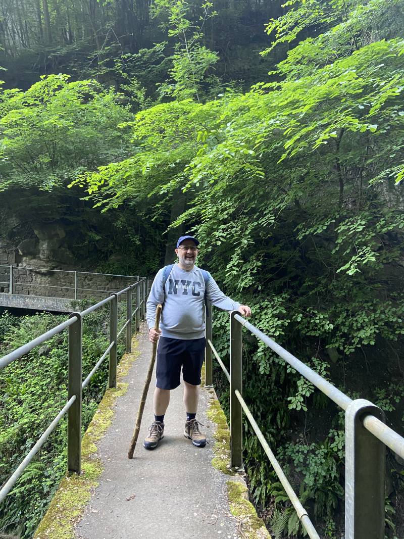 JF Poilprêt, souriant, arrêté sur une passerelle, tenant la rambarde de la main gauche et un bâton de la main droite, avec beaucoup d’arbres en arrière-plan.