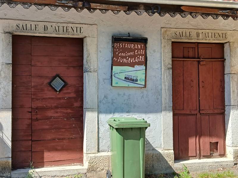 Façade crépie avec deux portes dont une condamnée et l’autre verrouillée, “Salle d’attente” inscrit au dessus de chacune ; entre les deux portes, une pancarte au mur, indiquant “Restaurant de l’ancienne gare du funiculaire”, une poubelle verte en dessous.