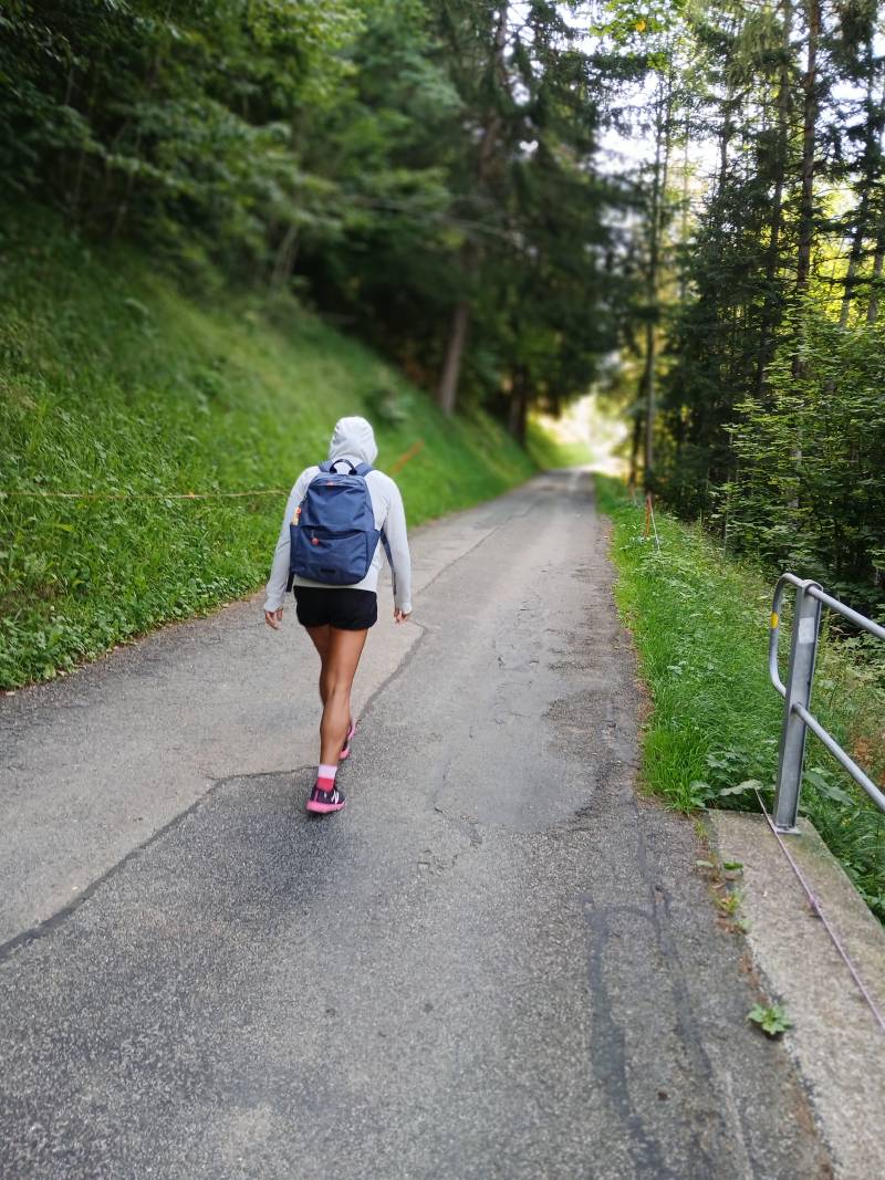 Mon épouse, de dos, avec coupe-vent gris et capuche, sac à dos bleu, en train de marcher sur la route goudronnée au départ de Haut de Caux