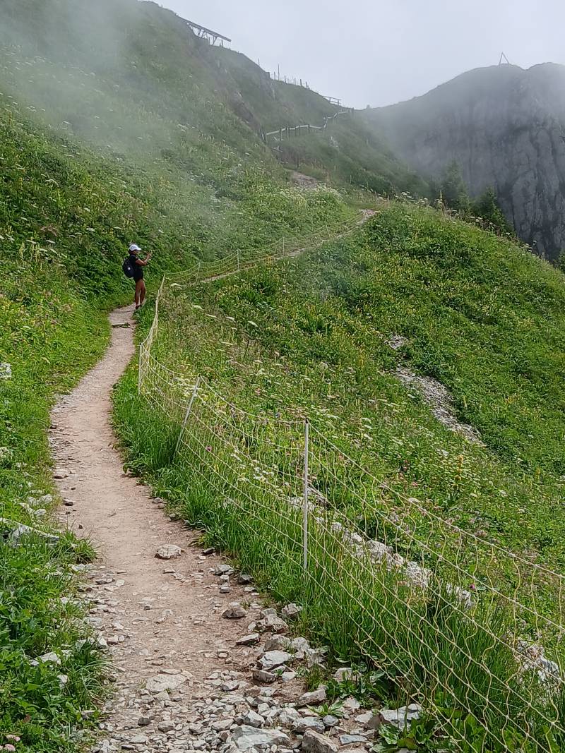 Montée sur un petit chemin de terre, la montagne verdoyante sur la gauche et la pente à droite, avec un petit filet pour éviter de tomber. Le chemin un peu raide zigzague plusieurs fois avant le sommet qu’on aperçoit au bout. La nappe de nuages s’éclaircit.