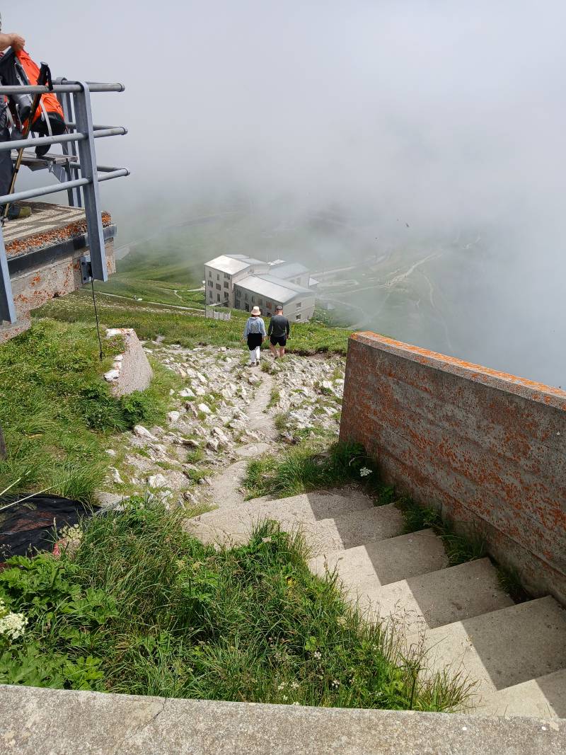 Vue sur un chemin de grosses pierres blanches menant à des escaliers. En bas, on aperçoit le bâtiment englobant le restaurant et la gare. Depuis le haut des escaliers, au même niveau, on voit une petite partie de la plateforme d’observation avec ses balustrades. Une épaisse couche de nuages cache une partie de la vue.