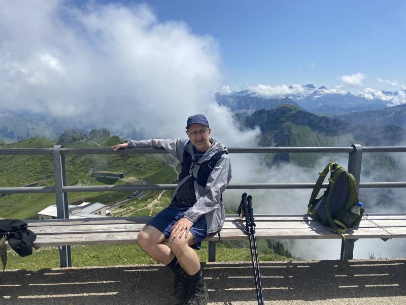 Moi, assis sur un banc de la plateforme, en arrière-plan sur la gauche, on voit la gare et un train qui s’approche, de l’autre côté on voit une petite partie des Alpes suisses; un nuage bouche la moitié de la vue.