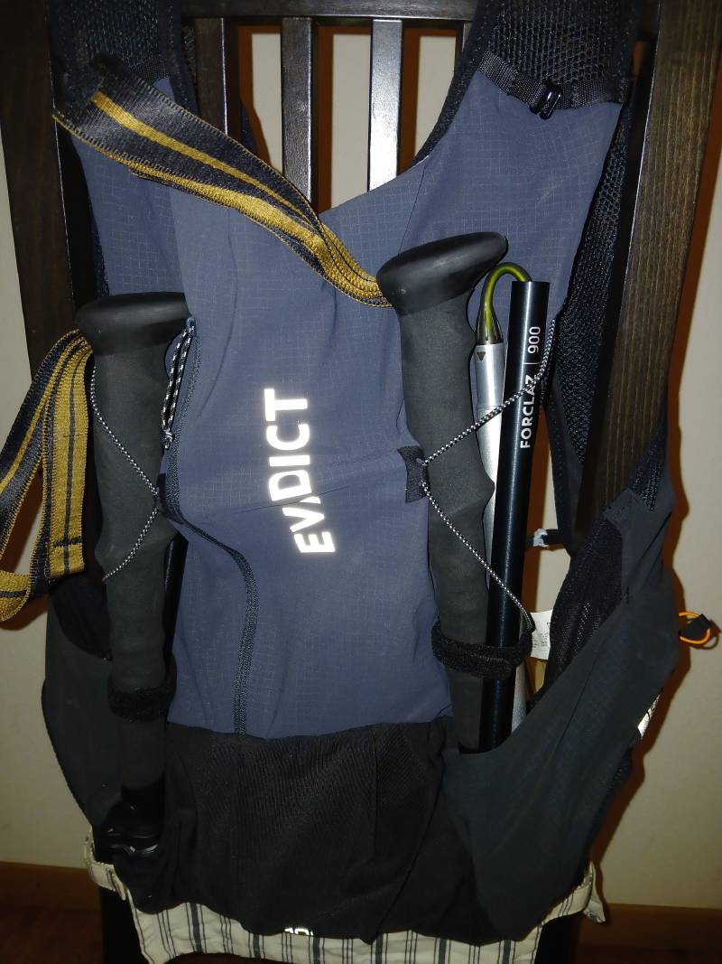 Sac de trail “EVADICT” bleu et noir, posé sur le dossier d’une chaise, avec de chaque côté un baton FORCLAZ 900 plié et retenu par un système d’élastiques.