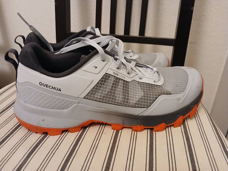 Une paire de chaussures de randonnées basses, blanches et grises, intérieur noir, semelles orange, posées sur une chaise. La marque QUECHUA apparaît, ainsi que le sigle “WEB-LOCK”